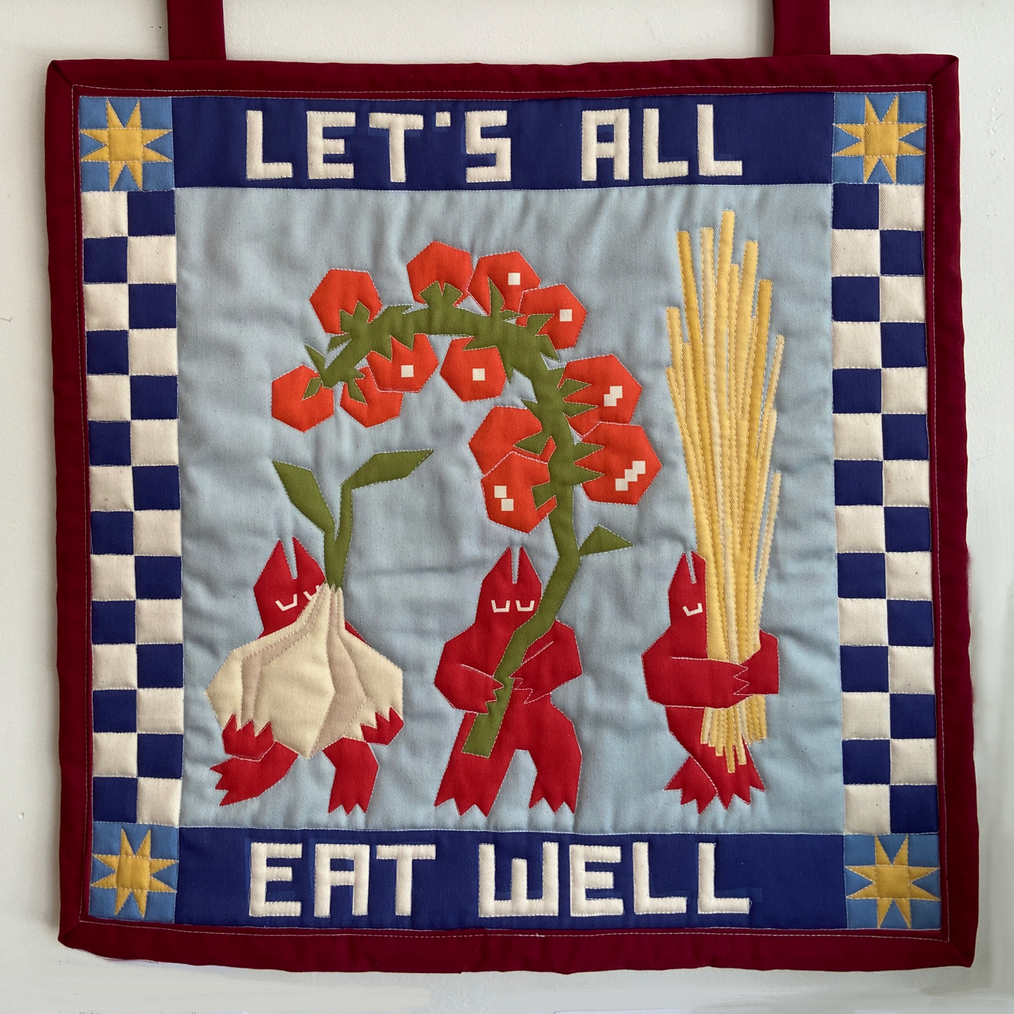 Quilt with tomato, garlic, and pasta design and 'Let's All Eat Well' text on a blue background